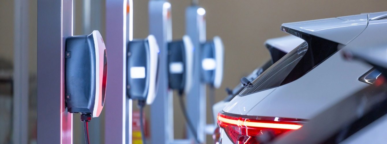 Reihe wandmontierter Ladestationen in einer Halle, rechts das Heck eines weißen Elektroautos mit leuchtendem Rücklicht
