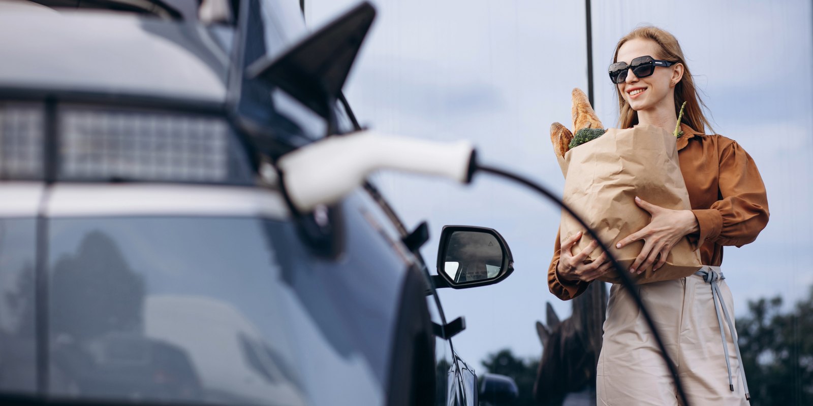 Frau mit Einkaufstüte neben einem ladenden Elektroauto